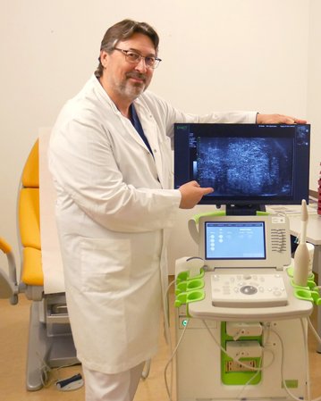 Ein Arzt in einem weißen Kittel steht neben einem Ultraschallgerät und zeigt auf den Monitor, der ein Ultraschallbild anzeigt. Das Gerät hat mehrere Bedienelemente und ist auf einem fahrbaren Wagen platziert.