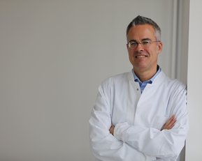 Ein Arzt in einem weißen Kittel steht lächelnd an einer Wand, die Arme verschränkt. Er trägt eine Brille und hat graue Haare.