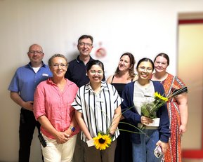 Eine Gruppe von sieben Personen steht zusammen, einige halten Blumen. Sie lächeln und posieren vor einer Wand mit einem roten Akzent.