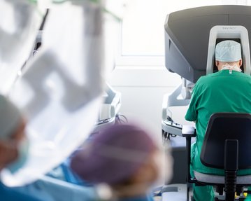 Ein Chirurg in grüner Kleidung sitzt vor einem Operationsroboter, während andere medizinische Fachkräfte im Hintergrund sichtbar sind.