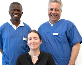 Drei Personen stehen und sitzen in einem medizinischen Umfeld. Zwei Männer in blauen Kitteln stehen hinter einer Frau in schwarzer Bluse, die auf einem Stuhl sitzt.