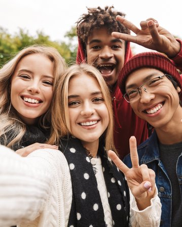 Vier junge Erwachsene posieren für ein Selfie, lächeln und zeigen Peace-Zeichen. Sie tragen lässige Kleidung und stehen in einer grünen Umgebung.