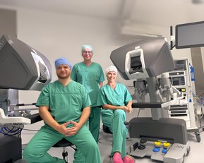 Drei medizinische Fachkräfte in grüner OP-Kleidung sitzen und stehen neben einem chirurgischen Robotersystem in einem Operationssaal.