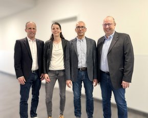 Vier Personen stehen in einem hellen Raum, lächelnd und in formeller Kleidung, bereit für ein Treffen oder eine Veranstaltung.