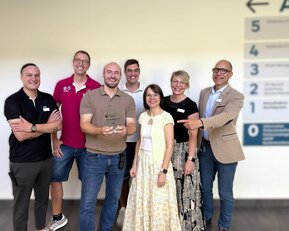 Eine Gruppe von sieben Personen steht zusammen, einige halten einen Preis. Im Hintergrund sind nummerierte Schilder sichtbar.