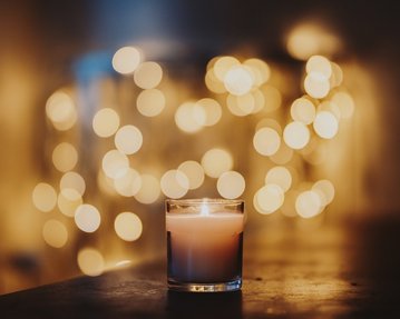 Eine brennende Kerze in einem klaren Glasbehälter steht im Vordergrund, während unscharfe, goldene Lichter im Hintergrund leuchten.
