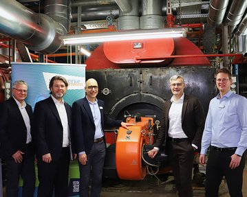 Fünf Personen stehen vor einem großen Kessel in einer Industrieumgebung, während sie an einem orangefarbenen Gerät arbeiten.