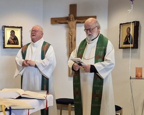 Zwei Priester in liturgischer Kleidung stehen in einer Kapelle, umgeben von religiösen Bildern und einem Kreuz. Ein Tisch mit einem offenen Buch ist im Vordergrund.