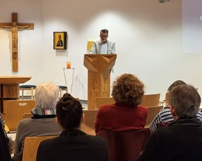 Ein Mann steht an einer Holzpult und spricht vor einer Gruppe von Zuhörern in einem Raum mit religiösen Symbolen.