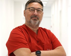 Ein Arzt in rotem Kittel steht mit verschränkten Armen in einem hellen Flur. Er trägt eine Brille und eine Smartwatch.