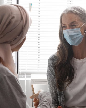 Zwei Frauen im Gespräch in einem Krankenhauszimmer. Eine trägt eine Kopfbedeckung und eine Maske, die andere hat graue Haare und eine Maske.