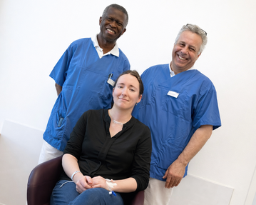 Drei Personen stehen und sitzen in einem modernen Raum. Zwei Männer in blauen Arbeitskleidung stehen hinter einer Frau, die in einem Stuhl sitzt.