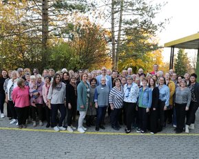 Eine große Gruppe von Menschen steht im Freien, umgeben von Bäumen, und posiert für ein Gruppenfoto. Es sind überwiegend Frauen zu sehen.