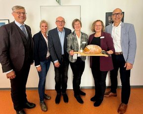 Sechs Personen stehen zusammen und halten ein großes Brot auf einem Tablett. Sie tragen formelle Kleidung und lächeln.